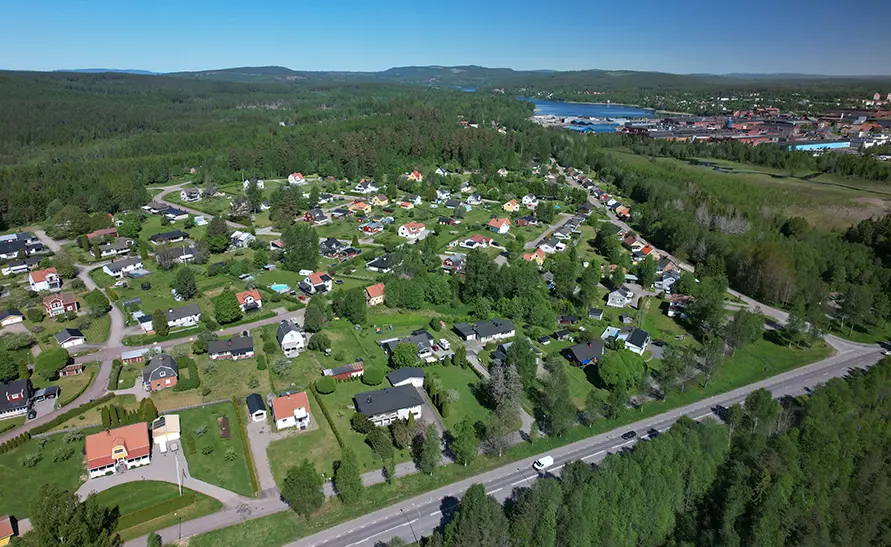 En vy över ett bostadsområde i Hagfors med villor och grönska, samt en sjö och bebyggelse i fjärran. En väg löper längs områdets kant.
